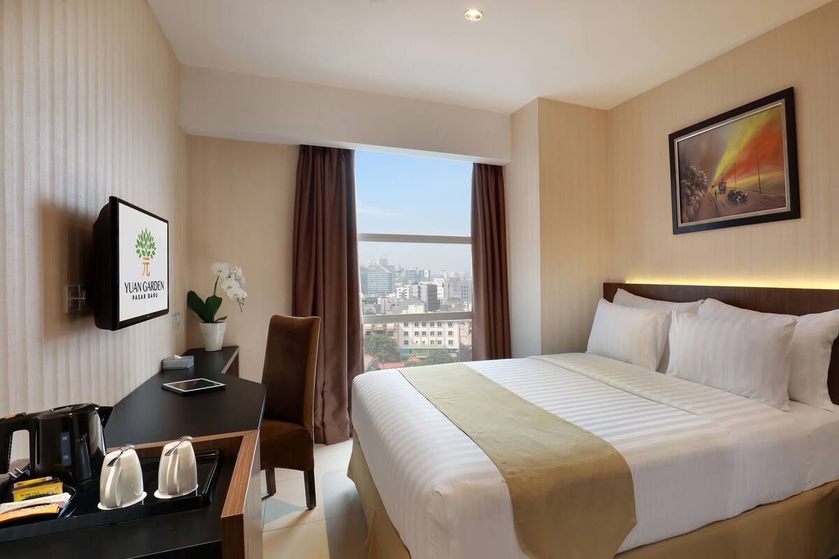A hotel room with a bed, a desk with a telephone, a chair, a window with curtains, and a painting on the wall.