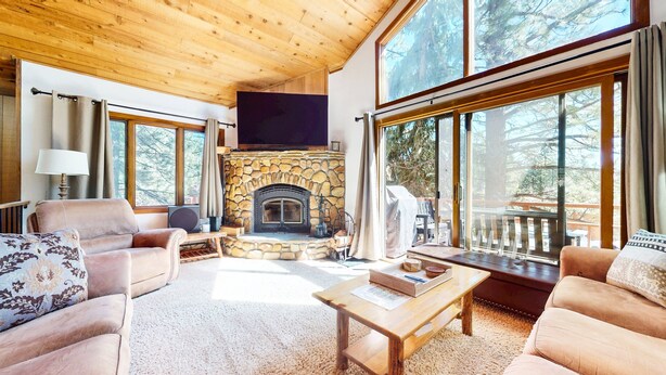 A cozy living room with a stone fireplace, large windows, and a wooden ceiling. - Woodlands 25 Updated and Spacious, Quiet with Forest Setting Views townhouse by RedAwning (Mammoth Lakes)