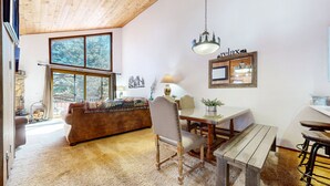 A cozy room with a wooden ceiling, a large window with a view of trees, a dining table with chairs, and a sofa with a blanket. - Woodlands 25 Updated and Spacious, Quiet with Forest Setting Views townhouse by RedAwning (Mammoth Lakes)
