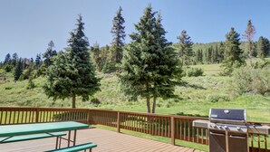 A wooden deck with a grill and picnic table, surrounded by a wooden fence and a view of a forested hillside.