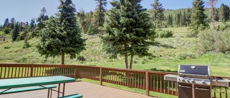 A wooden deck with a grill and picnic table, surrounded by a wooden fence and a view of a forested hillside.