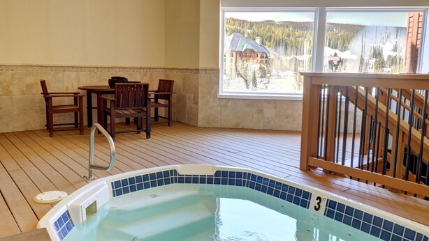A wooden deck with a hot tub, a dining area with chairs, and a view of a mountain landscape through large windows.