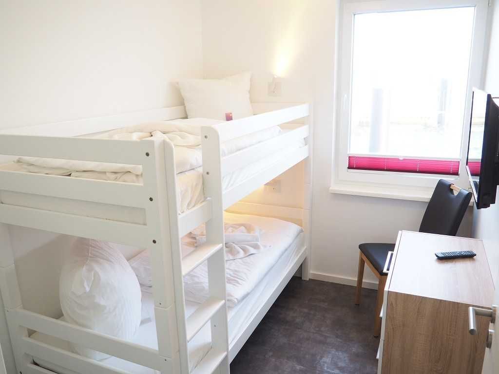A bunk bed room with a desk and chair, a window with blinds, and a television.