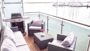 Ein Balkon mit Korbmöbeln, einem Grill und Blick auf Boote auf dem Wasser.