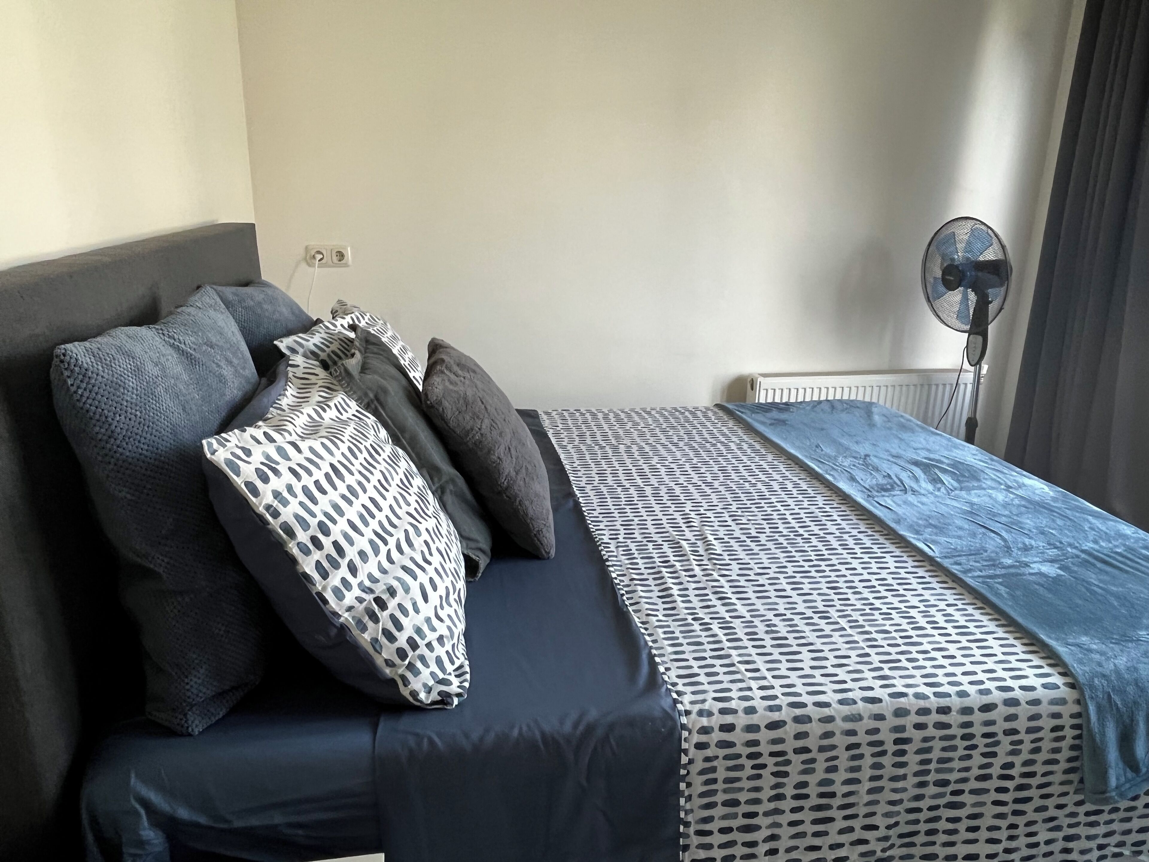 A neatly made bed with a patterned bedspread and pillows, a bedside table with a fan, and a radiator on the wall.