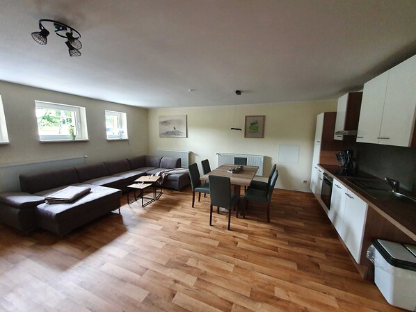 A modern living room with a sofa, dining table, and chairs. There is a kitchen area with cabinets and a countertop.