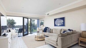 A spacious living room with a large window, a beige sofa, a round coffee table, and a white desk.