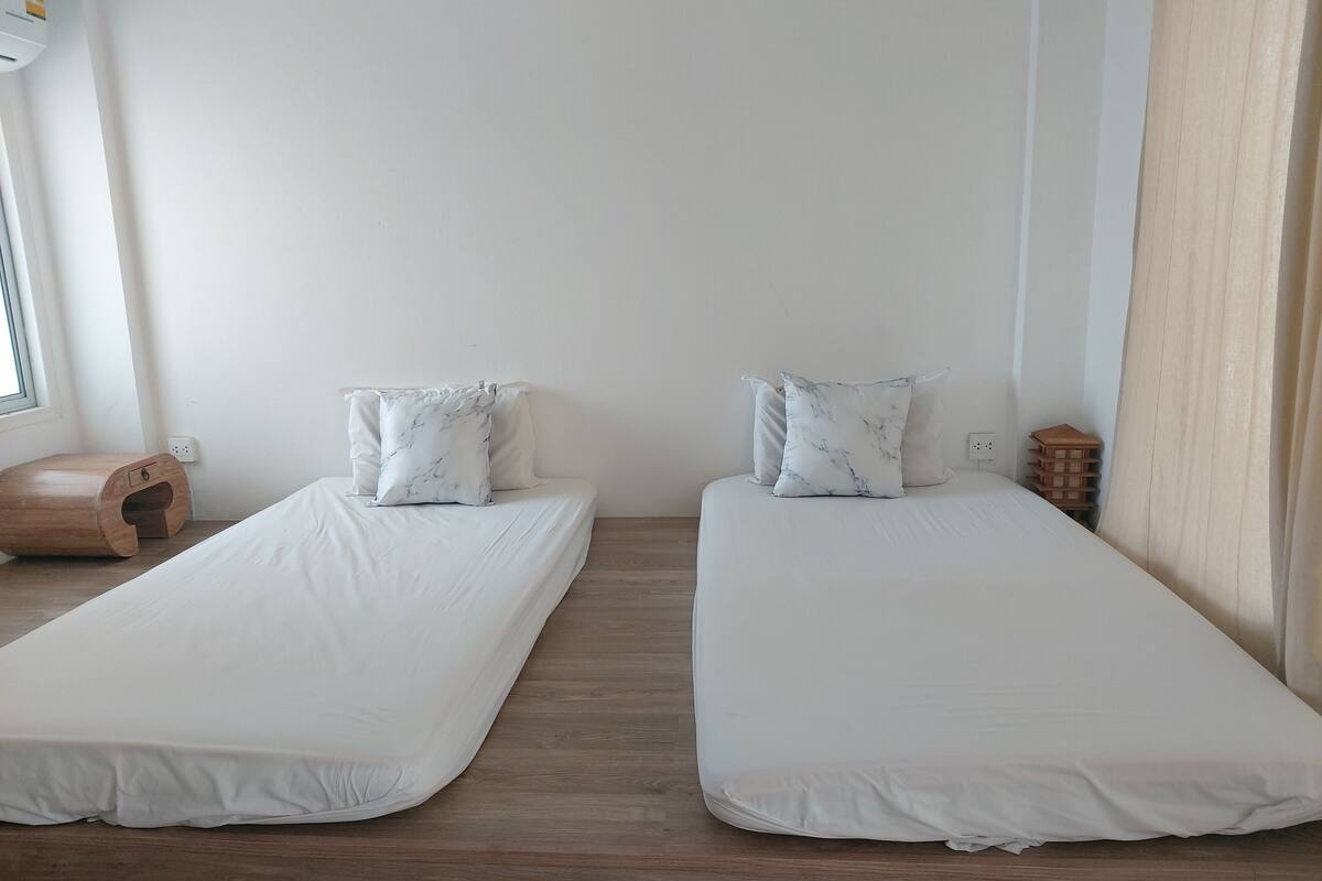 Two single beds with white bedding and marble-patterned pillows, wooden flooring, a small wooden stool, and a power outlet on the wall.