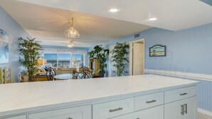 A spacious kitchen with a large island, white cabinetry, and a ceiling fan. There are potted plants and a sign on the wall. - Updated Condo Featuring Fresh Atlantic Breeze by RedAwning (Fernandina Beach)