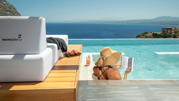Eine Person entspannt am Pool mit Blick auf das Meer, neben einem Laptop und einem Tablett mit Obst und Getränken.