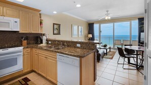 A kitchen with wooden cabinets, a microwave, and a dishwasher. There is a dining area with a table and chairs, and a view of the ocean through large windows. - 🏖️Beachfront, Fab Views-1BR+Bunk Boardwalk 805🏖️ (Panama City Beach)