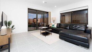 A modern living room with a black leather sofa, a coffee table, and a TV on a wooden stand. There is a sliding glass door leading to a patio area with outdoor furniture. - Resortstyle 4BR House With Parking@werribee (Werribee)