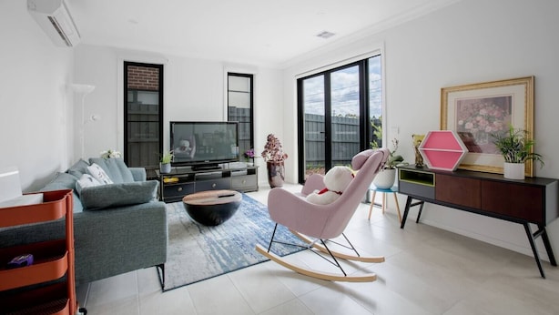 Un salon moderne avec un canapé gris, un fauteuil à bascule rose, un téléviseur à écran plat et une grande fenêtre donnant sur l’extérieur.