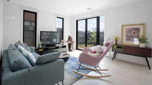Un salon moderne avec un canapé gris, un fauteuil à bascule rose, un téléviseur et une grande fenêtre donnant sur l’extérieur.