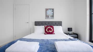 A neatly made bed with white and blue bedding, a grey headboard, and a decorative pillow. There is a framed picture on the wall, a bedside table with a lamp, and a window with blinds.