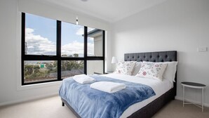 Une chambre moderne avec une grande fenêtre, un lit avec une literie bleue et une tête de lit rembourrée de couleur grise.