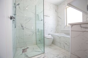 A modern bathroom with a glass-enclosed shower, white marble tiles, and a window providing natural light.