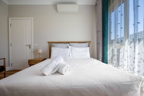 A neatly made bed with white linens and a rolled towel, a bedside table with a lamp, and a window with patterned curtains.
