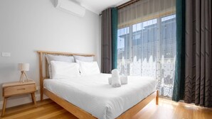 Ein ordentlich bezogenes Bett in einem Hotelzimmer, mit einem hölzernen Kopfteil, einem Nachttisch mit Lampe und einem Fenster mit Vorhängen.