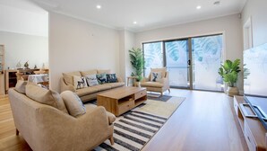 Ein modernes Wohnzimmer mit einer Couch, Sesseln, einem Couchtisch und einem Fernseher. Im Hintergrund befindet sich ein Essbereich mit Tisch und Stühlen.