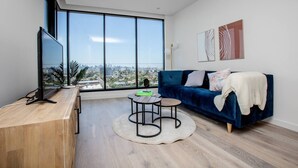 A modern living room with a large window, a blue sofa, a wooden desk, and a flat-screen TV. - The Lively City 2bed 2 Bath APT @footscray (Footscray)