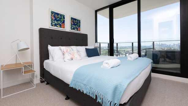 A modern bedroom with a large bed, a side table, and a view of the city through floor-to-ceiling windows. - The Lively City 2bed 2 Bath APT @footscray (Footscray)