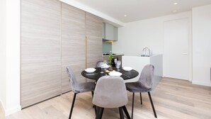 A modern dining area with a round table, grey upholstered chairs, and a minimalist kitchen in the background. - The Lively City 2bed 2 Bath APT @footscray (Footscray)
