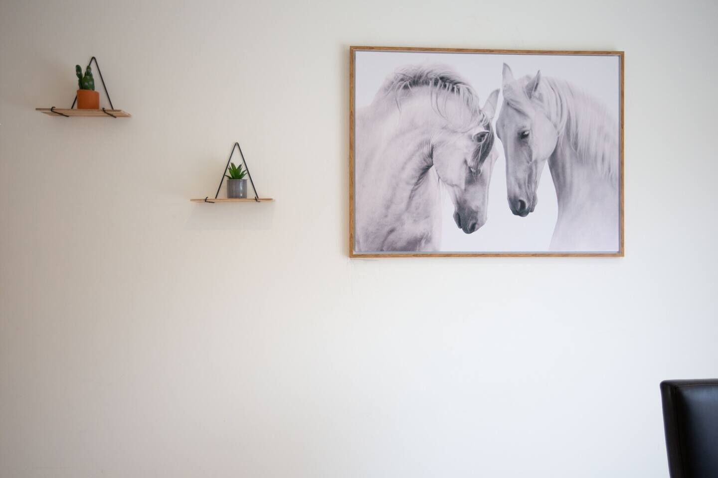 Una pared con dos estantes pequeños, cada uno con una planta y un objeto geométrico. También se ve un cuadro enmarcado de dos caballos.