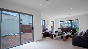 A modern living room with a sliding glass door leading to a wooden deck, a sofa set, and a coffee table. - Superb Luxe 5BR House@point Cook Near Lake (Point Cook)