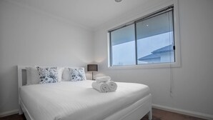 A neatly made bed with white bedding and floral-patterned pillows, a rolled towel, and a bedside lamp. - Superb Luxe 5BR House@point Cook Near Lake (Point Cook)