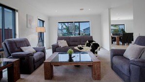 A modern living room with two sofas, a coffee table, and a dining area visible through large windows. - Superb Luxe 5BR House@point Cook Near Lake (Point Cook)