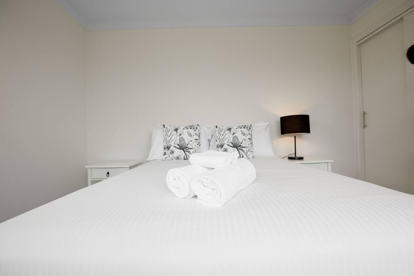 A neatly made bed with rolled towels, a black lamp on a bedside table, and floral-patterned pillows.