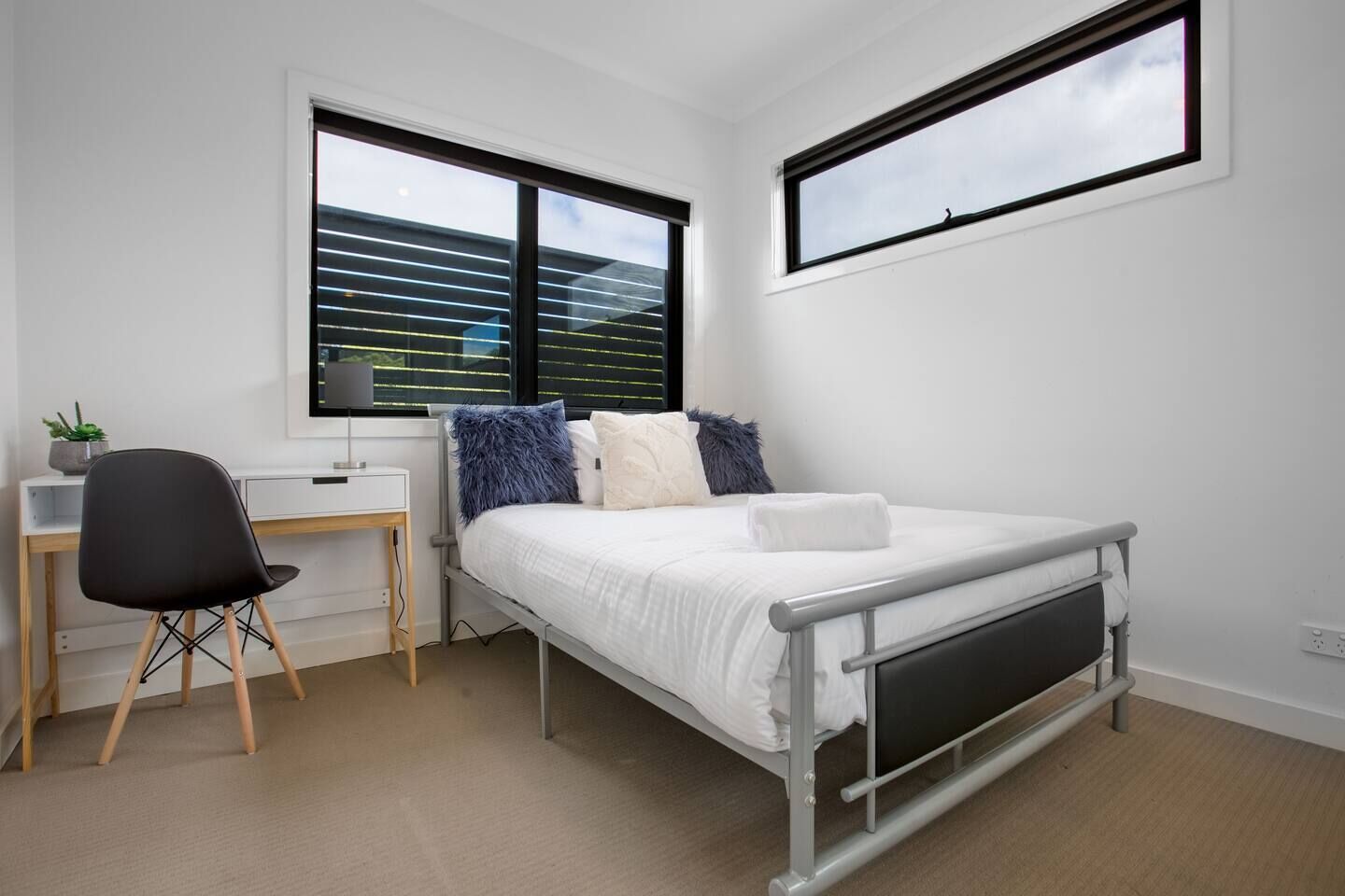 A bedroom with a bed, a desk, and a chair. There are two windows, one with blinds and one without.