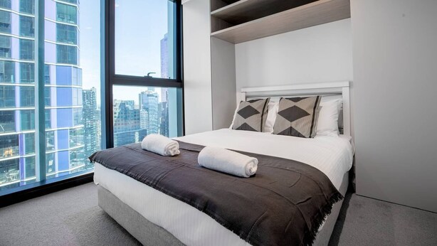A modern hotel room with a large window, a bed with a dark brown bedspread, and a city view. - Cozy APT With City Views Next to Meblourne Central (Melbourne)
