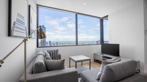 A modern living room with a sofa, a coffee table, and a TV. There is a large window with a city view. - Cozy APT Seaview Next to Southern Cross/pool/gym (Melbourne)