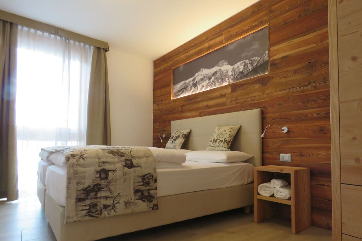 A bedroom with a wooden headboard, a bed with a patterned blanket, two pillows, a nightstand with a folded towel, and a framed picture on the wall.