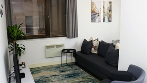 A modern living room with a dark sofa, a small side table, a radiator, a potted plant, and a window with blinds.