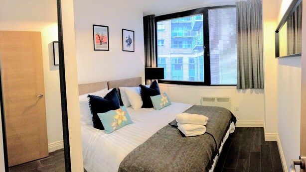 A hotel room with a large bed, a window with blinds, a door, and two framed pictures on the wall.