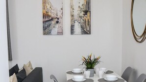 A dining area with a white table, grey chairs, and a grey sofa. There are two paintings on the wall, a ceiling light fixture, and a mirror on the right wall.
