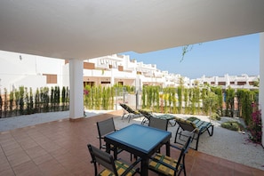 A covered outdoor patio with a table and chairs, surrounded by greenery and white buildings in the background. - Apartamento Rosa Chenz (Mar de Pulpi) (San Juan de los Terreros)