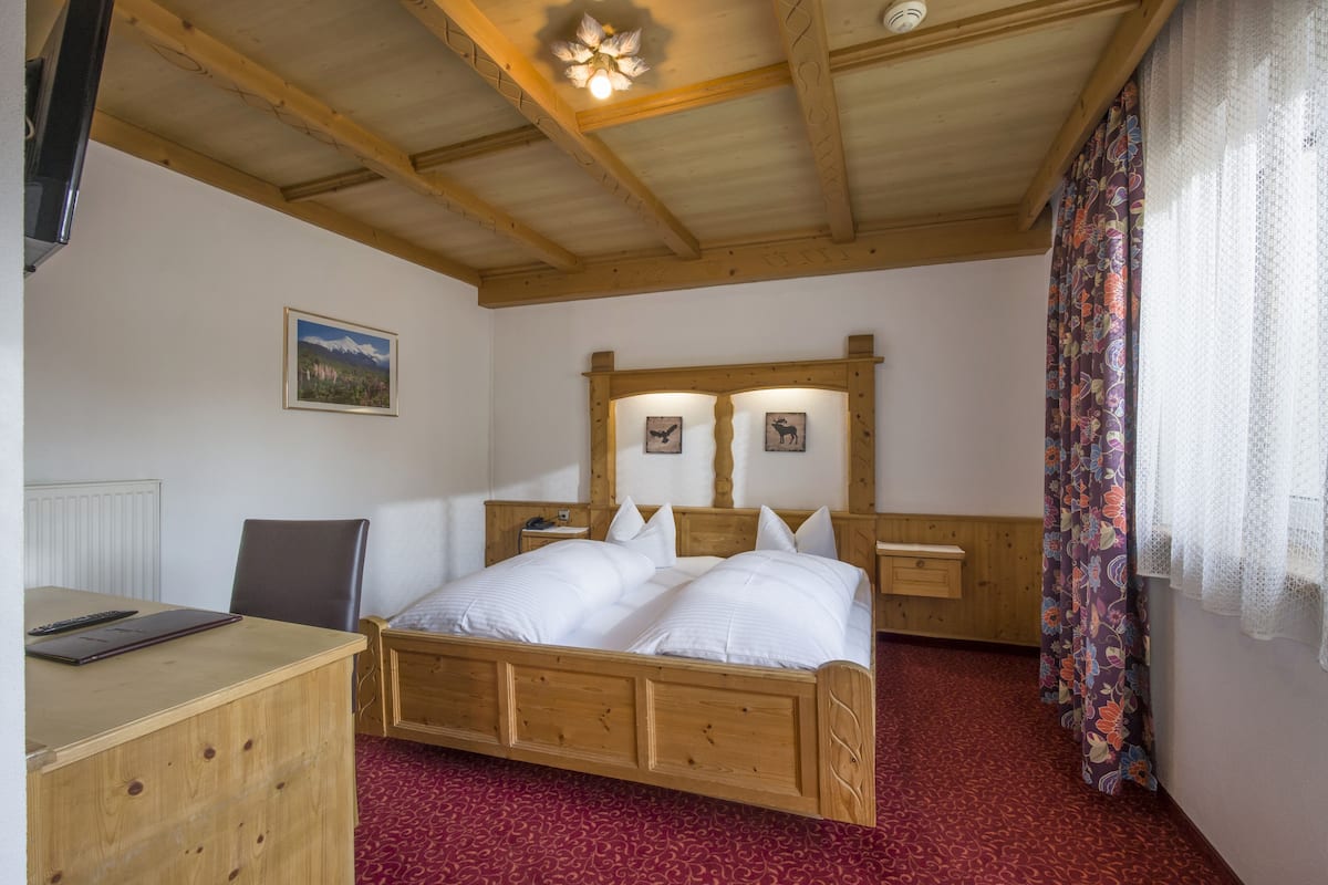 Ein Schlafzimmer mit einem hölzernen Bett, einem Schreibtisch mit einem Laptop, einem Stuhl, einem Nachttisch und einem Fenster mit Vorhängen.