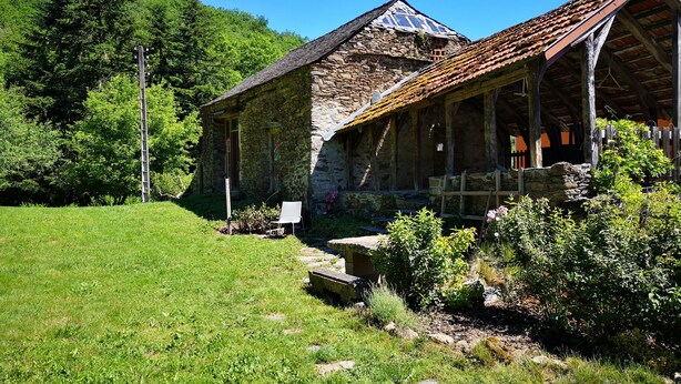 Un bâtiment en pierre doté d’un porche en bois, entouré d’une végétation luxuriante et surplombé d ’un ciel bleu azur.