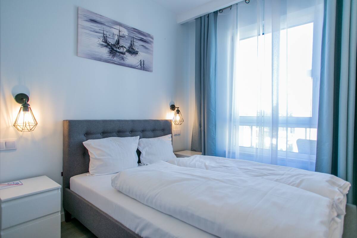 A neatly made bed with white linens, a grey upholstered headboard, and two wall-mounted reading lamps.