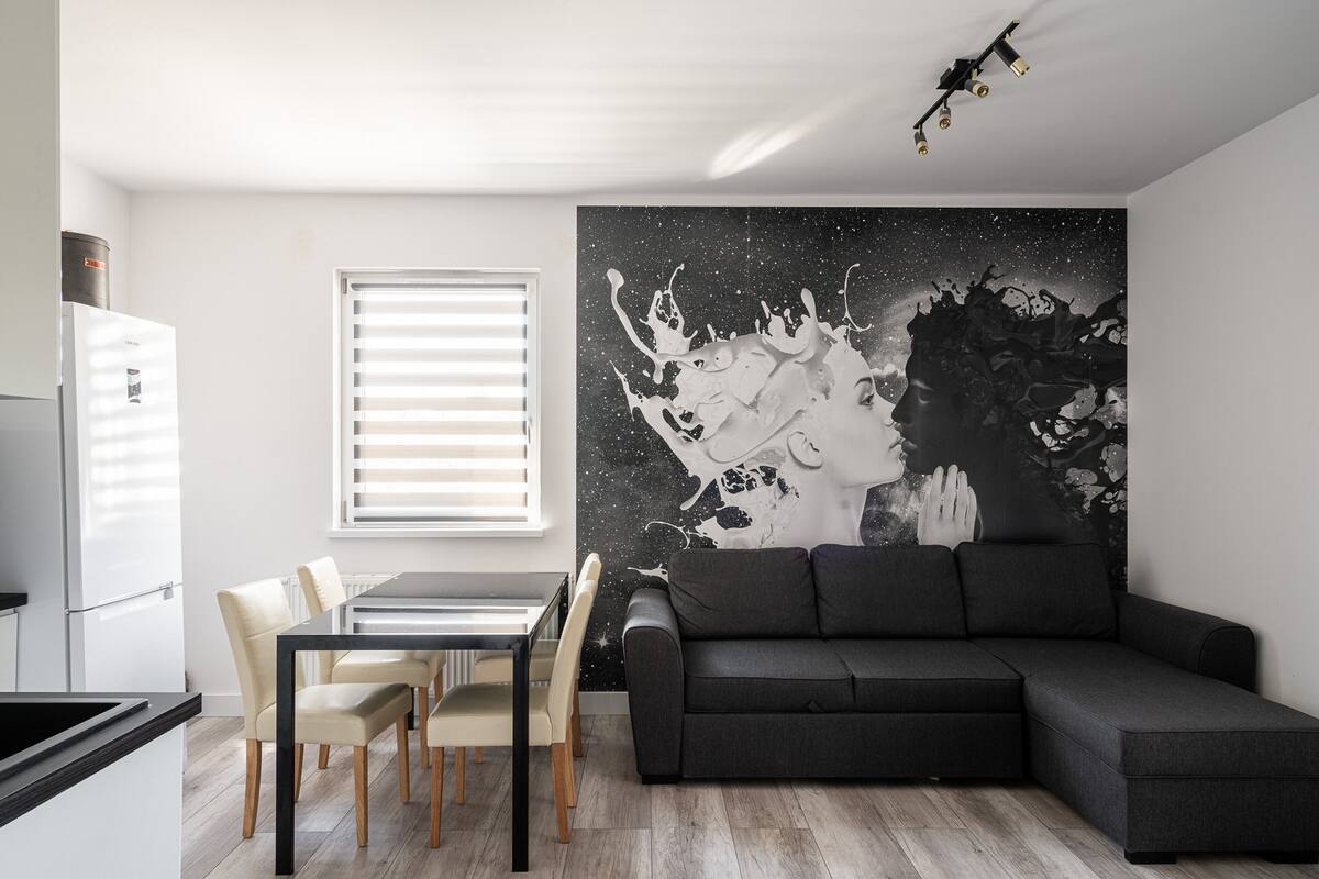 A modern living area with a dining table, chairs, a sofa, a kitchen with a refrigerator, and a large abstract wall art piece.