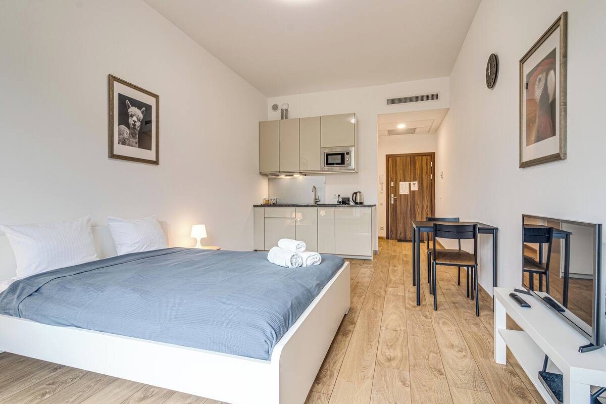 A modern hotel room with a bed, a small kitchenette, a dining area, and a TV.