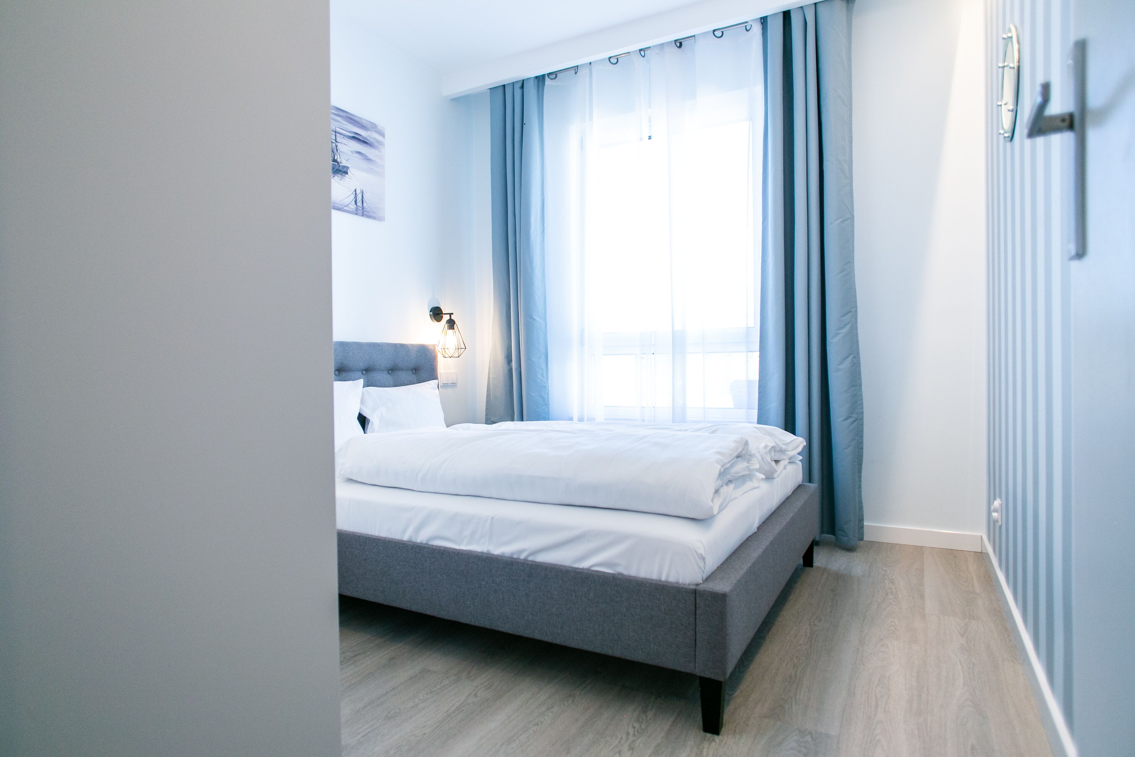 A modern bedroom with a grey bed, white bedding, a headboard, a bedside lamp, and a window with blue curtains.