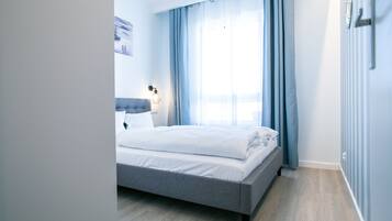 A modern bedroom with a grey bed, white bedding, a headboard, a bedside lamp, and a window with blue curtains.