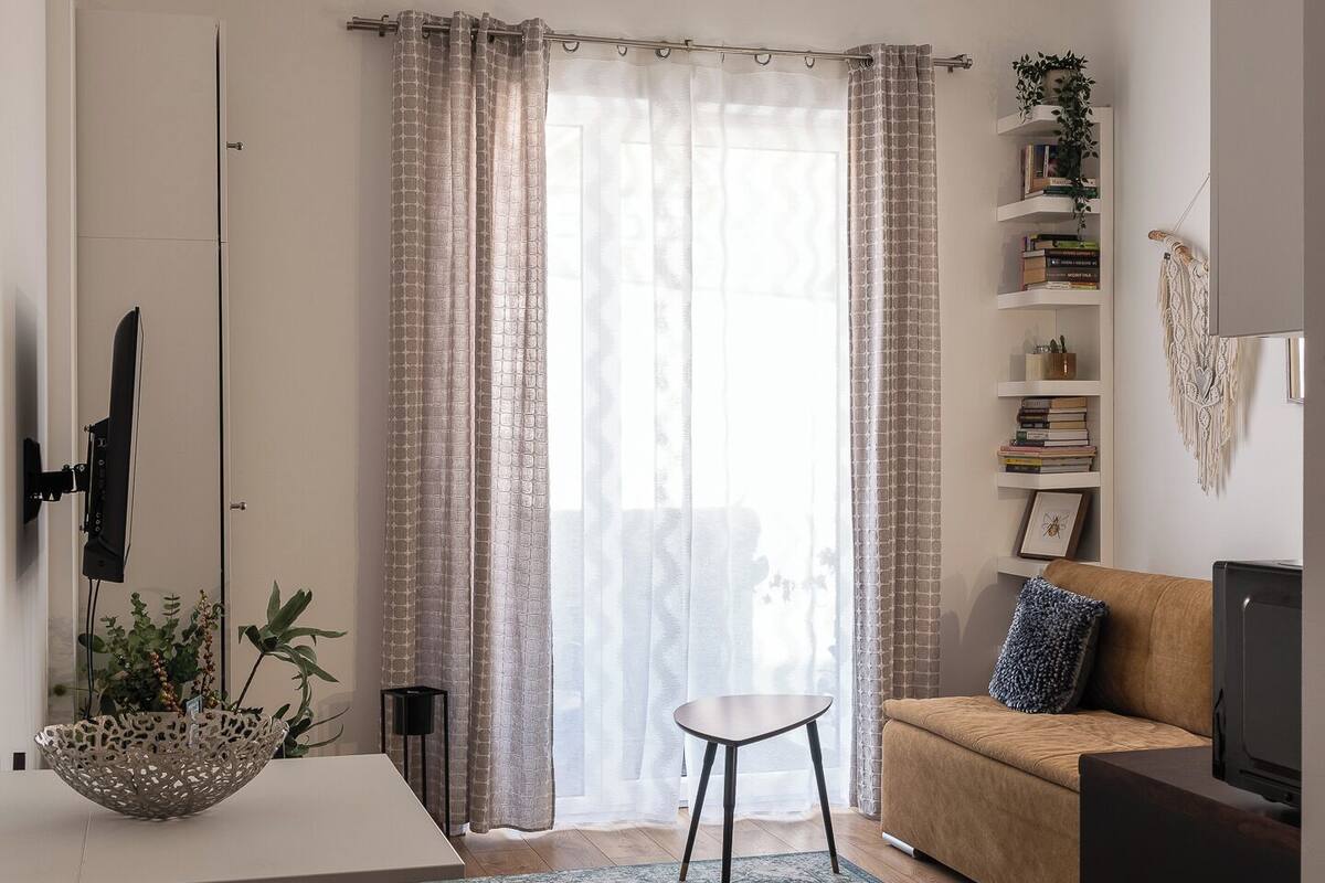 A modern living room with a wooden floor, a white table, a black chair, a beige sofa, and a TV. There is a bookshelf with books and a plant, and a window with curtains.