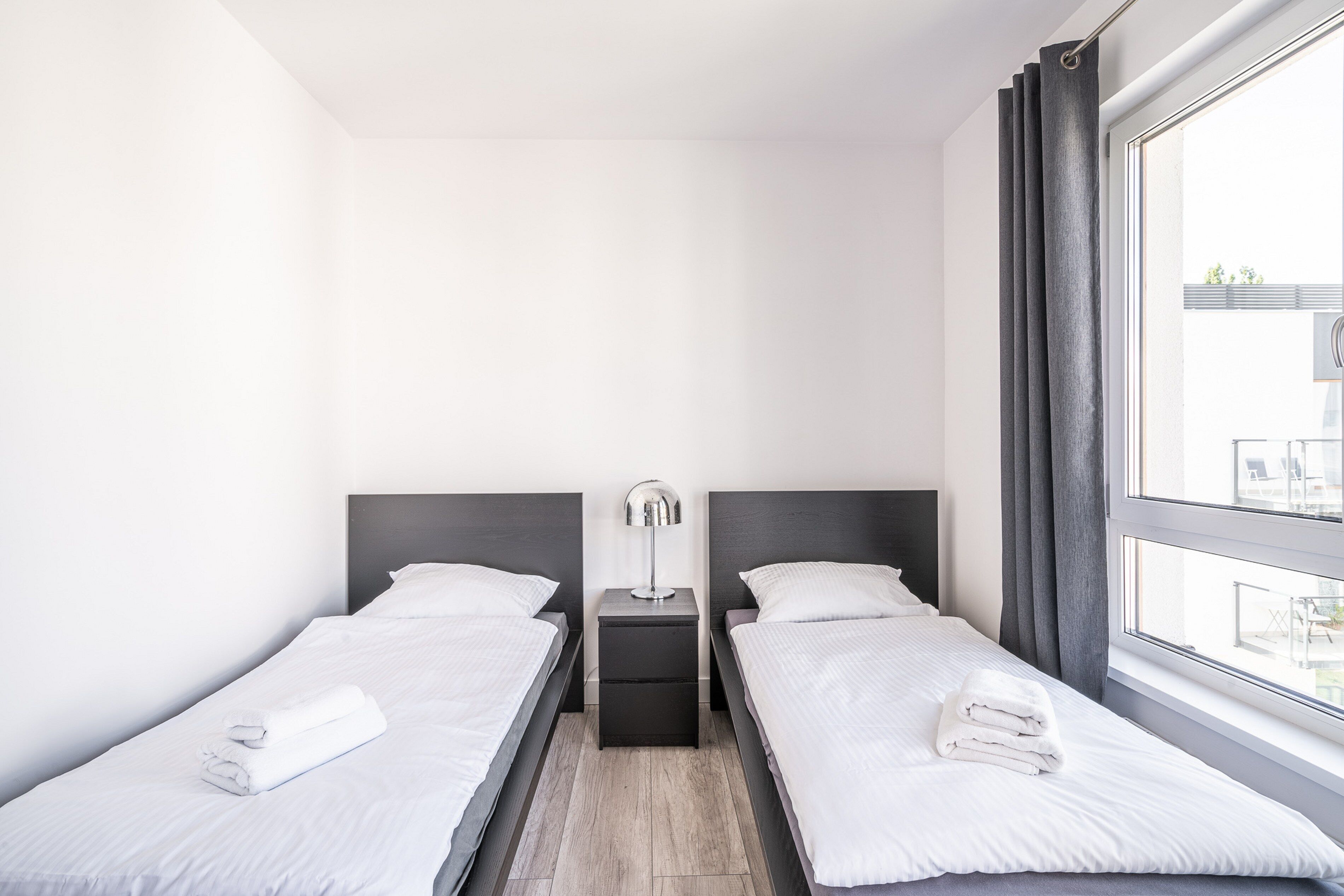 Une chambre d’hôtel moderne avec deux lits simples, une table de chevet avec une lampe et une grande fenêtre avec des rideaux.