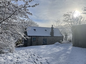 Uma casa de pedra com telhado coberto de neve e chaminé, rodeada por árvores carregadas de neve e um céu claro.